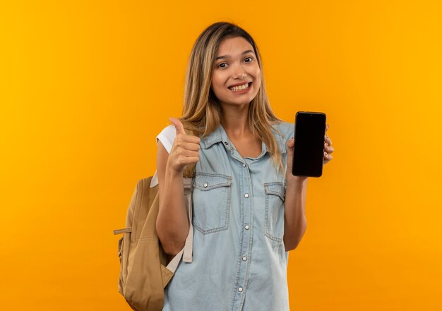Uśmiechnięta młoda ładna dziewczyna studentka na sobie plecak pokazujący telefon komórkowy i kciuk na białym tle na pomarańczowej ścianie