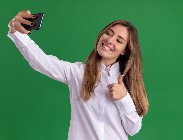 Uśmiechnięta młoda ładna kaukaska dziewczyna kciuki w górę, trzymająca i patrząca na telefon, biorąc selfie