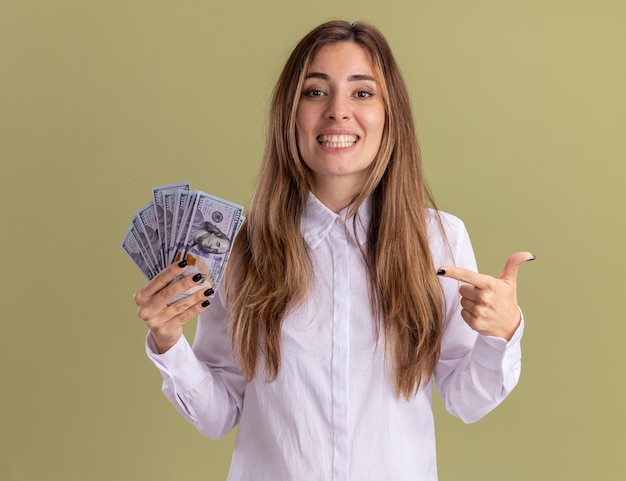 Uśmiechnięta młoda ładna kaukaska dziewczyna trzyma i wskazuje pieniądze odizolowane na oliwkowozielonej ścianie z miejscem na kopię