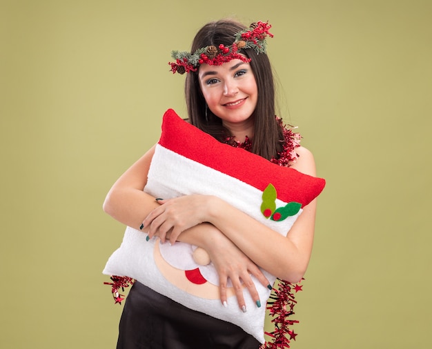 Uśmiechnięta młoda ładna kaukaska dziewczyna ubrana w świąteczny wieniec z głowy i blichtrową girlandę na szyi trzymającą poduszkę świętego mikołaja patrząc na kamerę odizolowaną na oliwkowozielonym tle z kopią przestrzeni