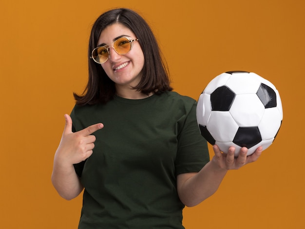 Uśmiechnięta młoda ładna kaukaska dziewczyna w okularach przeciwsłonecznych trzymająca i wskazująca na piłkę