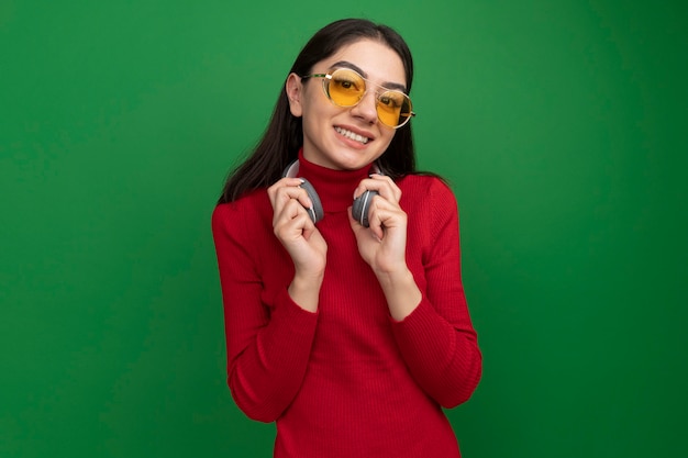 Uśmiechnięta młoda ładna kaukaska kobieta w okularach przeciwsłonecznych i słuchawkach na szyi chwytających słuchawki
