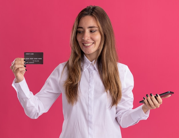 Uśmiechnięta młoda ładna kaukaski dziewczyna trzyma telefon i patrzy na kartę kredytową na białym tle na różowej ścianie z miejsca na kopię