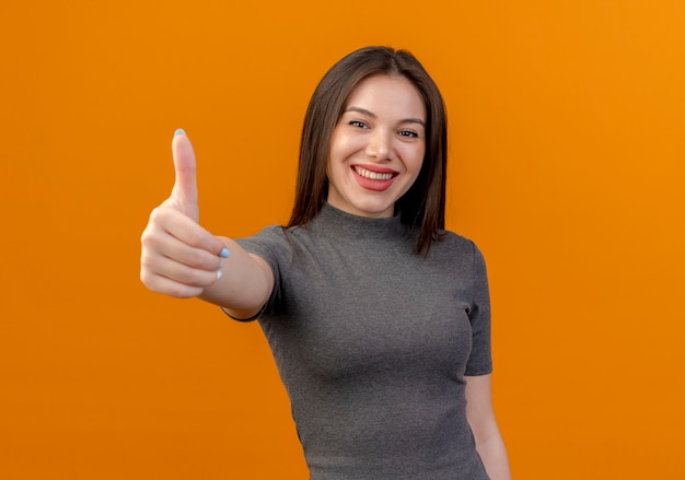 Uśmiechnięta młoda ładna kobieta pokazuje kciuk do góry na białym tle na pomarańczowym tle z miejsca na kopię