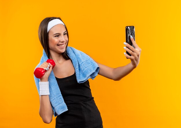 Uśmiechnięta młoda ładna sportowa dziewczyna z opaską na głowę i opaską, trzymając i patrząc na telefon komórkowy i trzymając hantle z ręcznikiem na szyi na białym tle na pomarańczowej przestrzeni