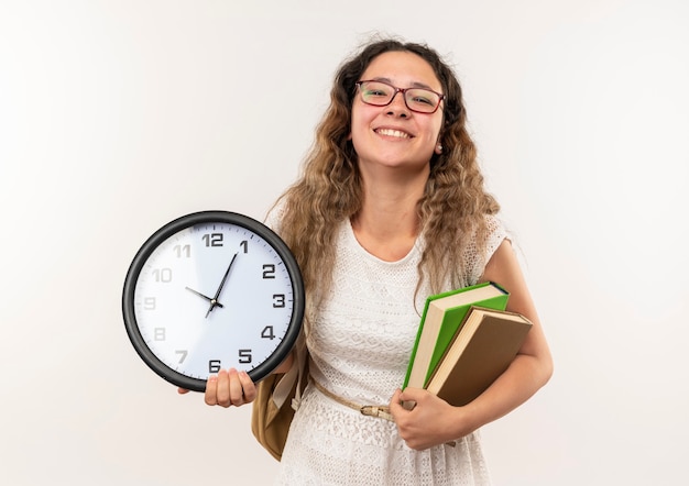 Uśmiechnięta młoda ładna uczennica w okularach i plecak trzymając książki i zegar na białym tle na białej ścianie