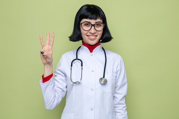 Uśmiechnięta młoda ładna w okularach optycznych kaukaska dziewczyna w mundurze lekarza ze stetoskopem, gestykulująca trzy palcami