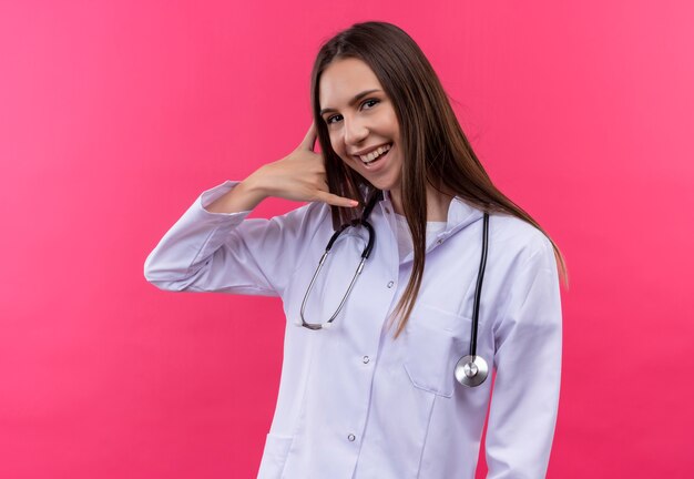 Uśmiechnięta młoda lekarka dziewczyna ubrana stetoskop medycznej sukni pokazujący gest połączenia telefonicznego na na białym tle różowym tle