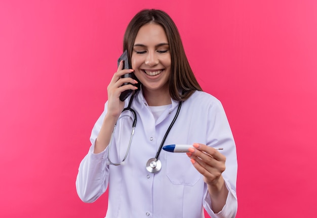 Uśmiechnięta młoda lekarka dziewczyna ubrana w stetoskop medycznej sukni mówi przez telefon, patrząc na termometr om jej rękę na na białym tle różowym tle