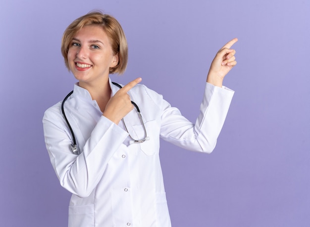 Uśmiechnięta młoda lekarka ubrana w szatę medyczną ze stetoskopem wskazuje z boku na białym tle na niebieskim tle z miejscem na kopię