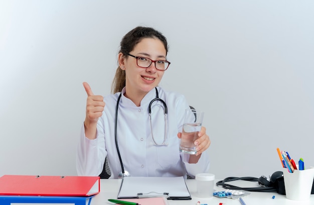 Uśmiechnięta młoda lekarka w szlafroku medycznym i stetoskopie i okularach siedzi przy biurku z narzędziami medycznymi patrząc trzymając szklankę wody pokazując kciuk w górę na białym tle