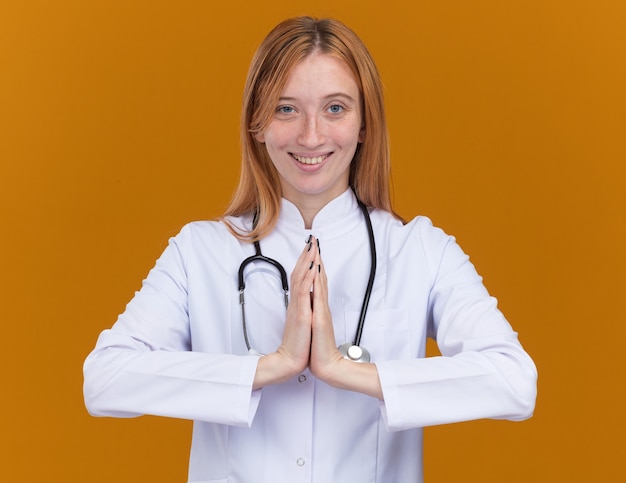 Bezpłatne zdjęcie uśmiechnięta młoda lekarka z imbirem w szacie medycznej i stetoskopie trzymająca ręce razem