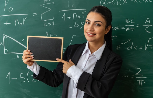 uśmiechnięta młoda nauczycielka stojąca przed tablicą trzymająca mini tablicę w klasie