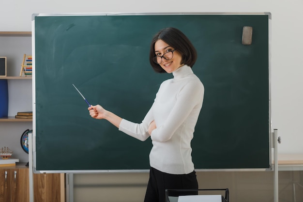 uśmiechnięta młoda nauczycielka w okularach stojąca przed tablicą i wskazująca na tablicę ze wskaźnikiem w klasie