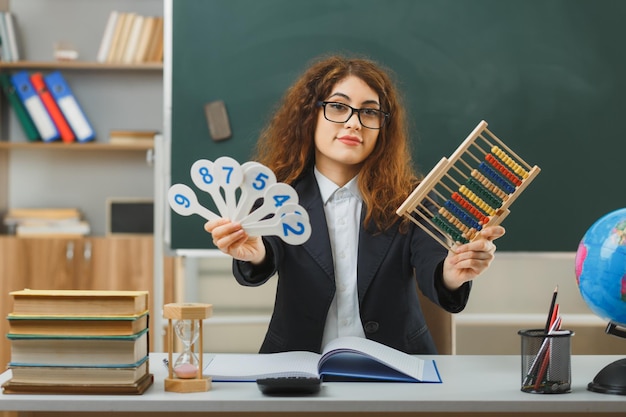 uśmiechnięta młoda nauczycielka w okularach trzymająca liczydło z wentylatorem numerycznym siedząca przy biurku z szkolnymi narzędziami w klasie