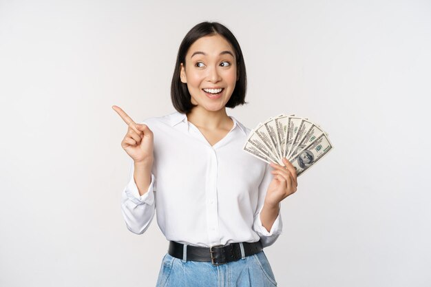 Uśmiechnięta młoda nowoczesna azjatycka kobieta wskazująca na baner reklamowy trzymająca pieniądze w gotówce stojące na białym tle