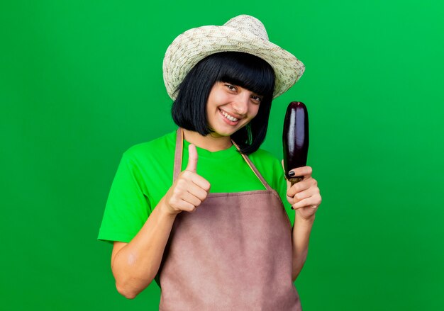 Uśmiechnięta młoda ogrodniczka w mundurze w kapeluszu ogrodniczym trzyma bakłażana