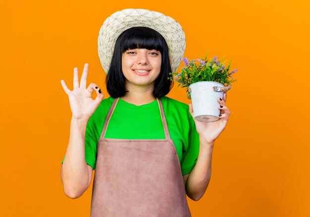 Uśmiechnięta młoda ogrodniczka w mundurze w kapeluszu ogrodniczym trzyma doniczkę
