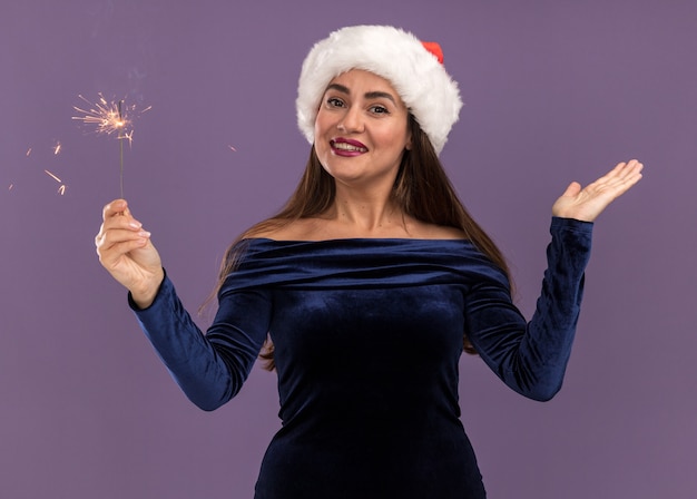 Uśmiechnięta młoda piękna dziewczyna ubrana w niebieską sukienkę i kapelusz Boże Narodzenie trzymając zimne ognie, rozkładając rękę na białym tle na fioletowym tle