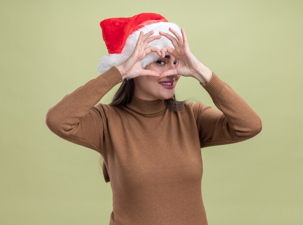 Uśmiechnięta młoda piękna dziewczyna ubrana w świąteczny kapelusz pokazujący gest serca na oliwkowym tle