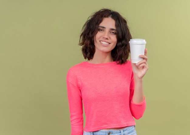 Uśmiechnięta młoda piękna kobieta trzyma plastikową filiżankę kawy na odosobnionej zielonej ścianie z miejsca na kopię