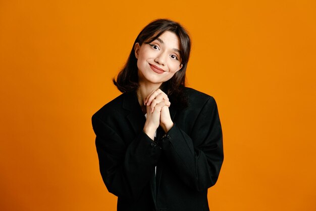 Uśmiechnięta młoda piękna kobieta ubrana w czarną kurtkę odizolowaną na pomarańczowym tle