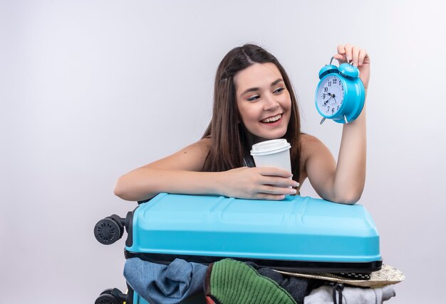 Uśmiechnięta młoda podróżna kobieta trzyma budzik i podróżuje plastikowy kubek kawy z walizką na odizolowanej białej ścianie