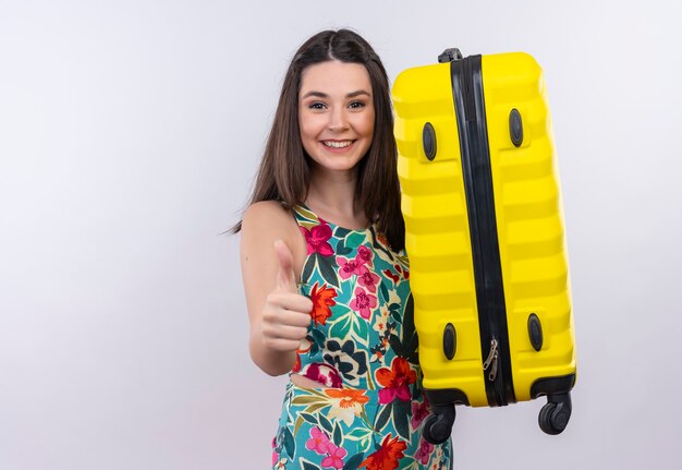 Uśmiechnięta młoda podróżniczka kobieta trzyma walizkę pokazując kciuk do góry na odosobnionej białej ścianie