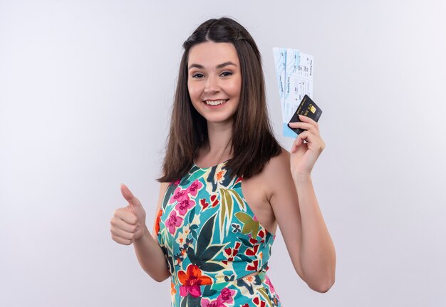 Uśmiechnięta młoda podróżniczka kobieta ubrana w wielokolorową sukienkę, trzymając bilety i kciuk na białej ścianie