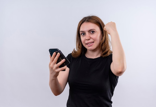 Uśmiechnięta młoda przypadkowa kobieta trzyma telefon komórkowy i podnosi pięść na odosobnionej białej przestrzeni z kopią miejsca