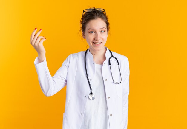 Uśmiechnięta młoda słowiańska dziewczyna w mundurze lekarza ze stetoskopem, udając, że trzyma coś odizolowanego na pomarańczowej ścianie z miejscem na kopię