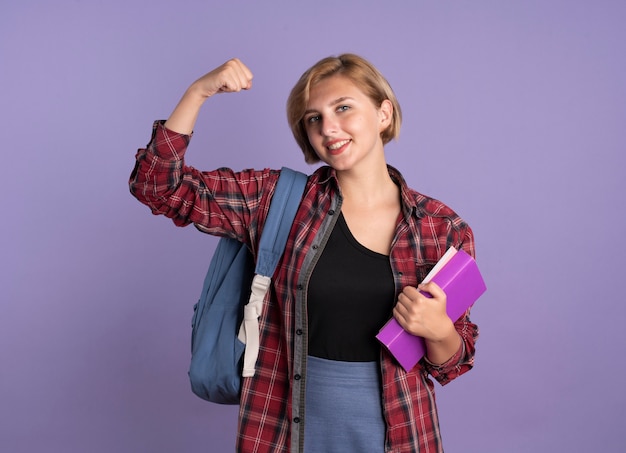 Uśmiechnięta Młoda Słowiańska Studentka Nosząca Plecak Napina Biceps, Trzyma Książkę I Notatnik