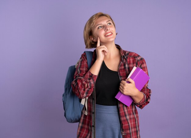 Uśmiechnięta młoda słowiańska studentka w plecaku kładzie palec na twarzy trzyma książkę i notatnik patrząc w górę