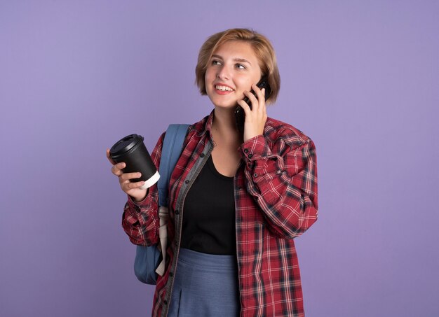 Uśmiechnięta młoda słowiańska studentka w plecaku trzyma papierowy kubek rozmawia przez telefon, patrząc na bok