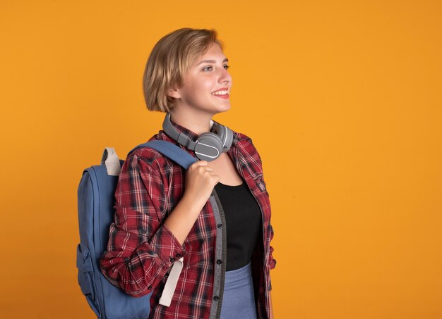 Uśmiechnięta młoda słowiańska studentka ze słuchawkami w plecaku stoi bokiem