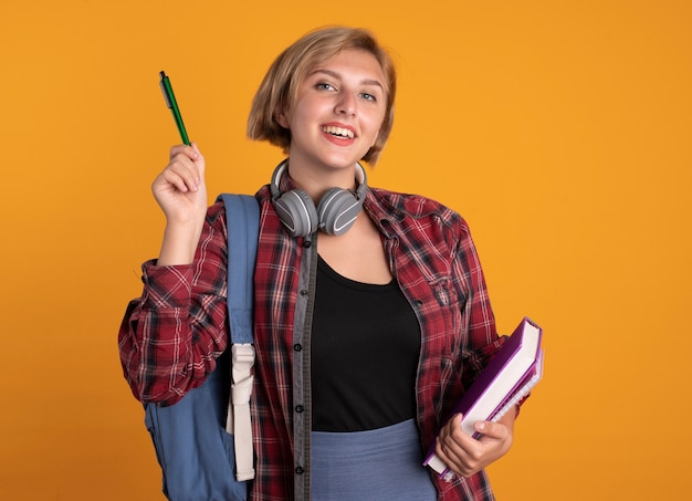 Uśmiechnięta młoda słowiańska studentka ze słuchawkami w plecaku trzyma długopis i notatnik