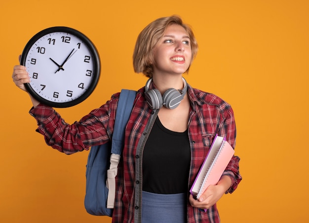 Bezpłatne zdjęcie uśmiechnięta młoda słowiańska studentka ze słuchawkami w plecaku trzyma książkę z zegarem i notatnik patrząc na bok at