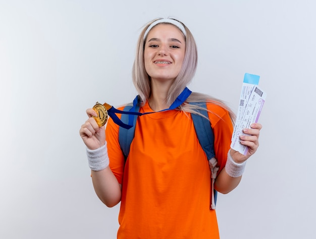 Uśmiechnięta młoda sportowa dziewczyna rasy kaukaskiej ze złotym medalem na szyi, nosząca opaskę na głowę z plecakiem i opaski na nadgarstki, trzyma bilety lotnicze