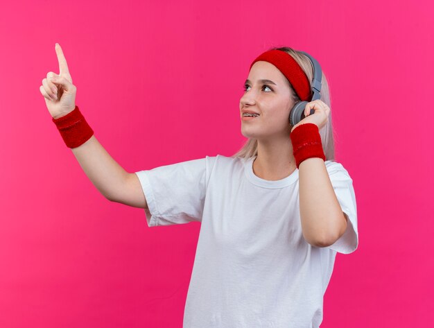 Uśmiechnięta młoda sportowa kobieta z szelkami na słuchawkach nosząca pałąk i opaski na rękę wygląda i wskazuje na bok na białym tle na różowej ścianie