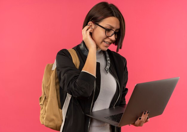Uśmiechnięta młoda studencka dziewczyna w okularach i plecak trzymając i patrząc na laptopa, trzymając rękę w pobliżu ucha na białym tle na różowej ścianie