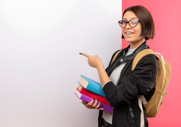 Uśmiechnięta Młoda Studentka W Okularach I Plecak Trzymając Książki Stojące Przed Białą ścianą, Wskazując Na To Na Białym Tle Na Różowej ścianie