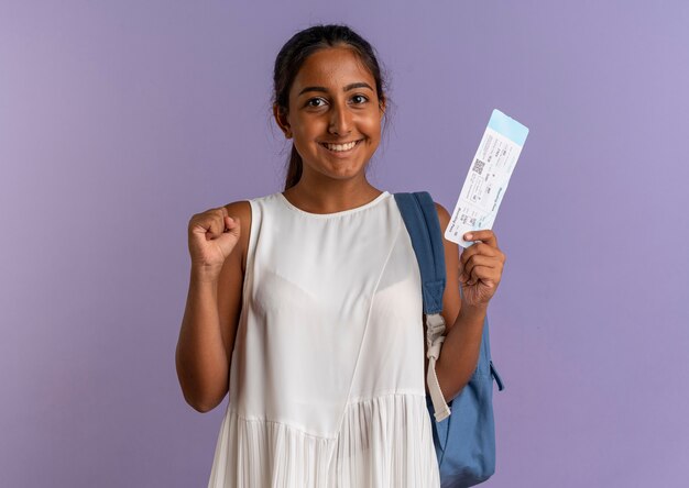 Uśmiechnięta młoda uczennica noszenie plecaka trzymając bilet i pokazując gest tak na na białym tle fioletowy