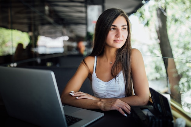 Uśmiechnięta modelka pracuje na swoim laptopie na tarasie kawiarni daytilme