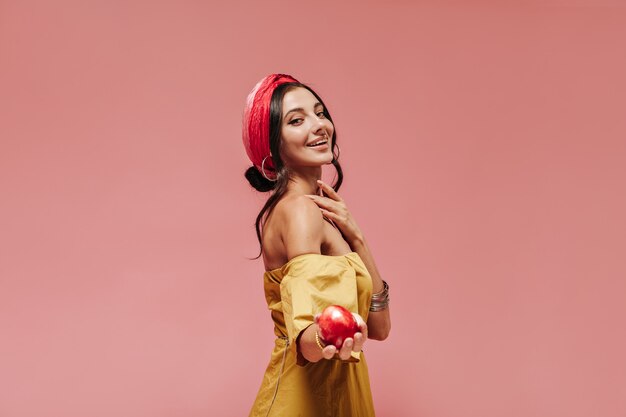 Uśmiechnięta modna dama z kręconymi włosami w czerwonej chustce, dodatkach i żółtej jasnej sukience, trzymająca czerwone jabłko i patrząca na przód