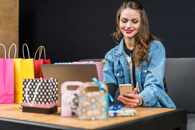 Uśmiechnięta modna kobieta siedzi w domu z torba na zakupy; laptop i smartfon