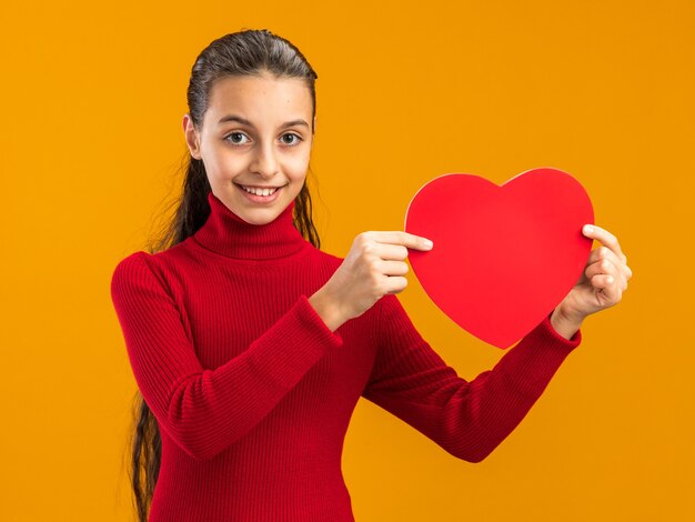 Uśmiechnięta nastolatka pokazująca kształt serca przed kamerą patrzącą na przód na pomarańczowej ścianie