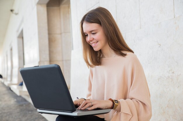 Uśmiechnięta nastoletnia dziewczyna wyszukuje na laptopie na ławce przy budynkiem