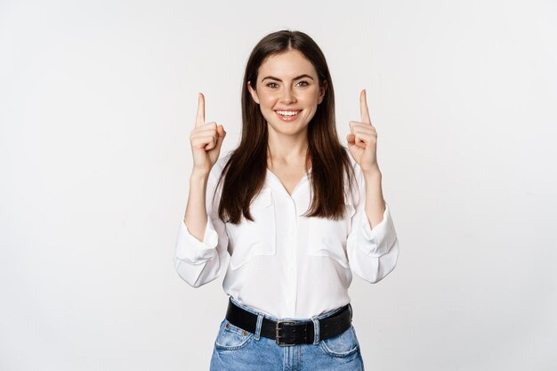 Uśmiechnięta pewna siebie ładna kobieta, wskazująca palcami w górę, pokazująca baner z logo, ogłoszenie, stojąca w białej bluzce na tle studia.