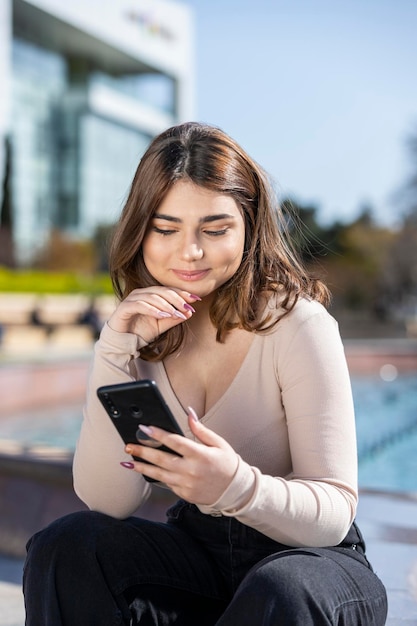 Uśmiechnięta piękna dziewczyna trzyma telefon i siedzi w parku Wysokiej jakości zdjęcie