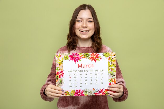 Uśmiechnięta piękna młoda dziewczyna na szczęśliwy dzień kobiet trzymająca kalendarz przed kamerą odizolowana na oliwkowozielonej ścianie